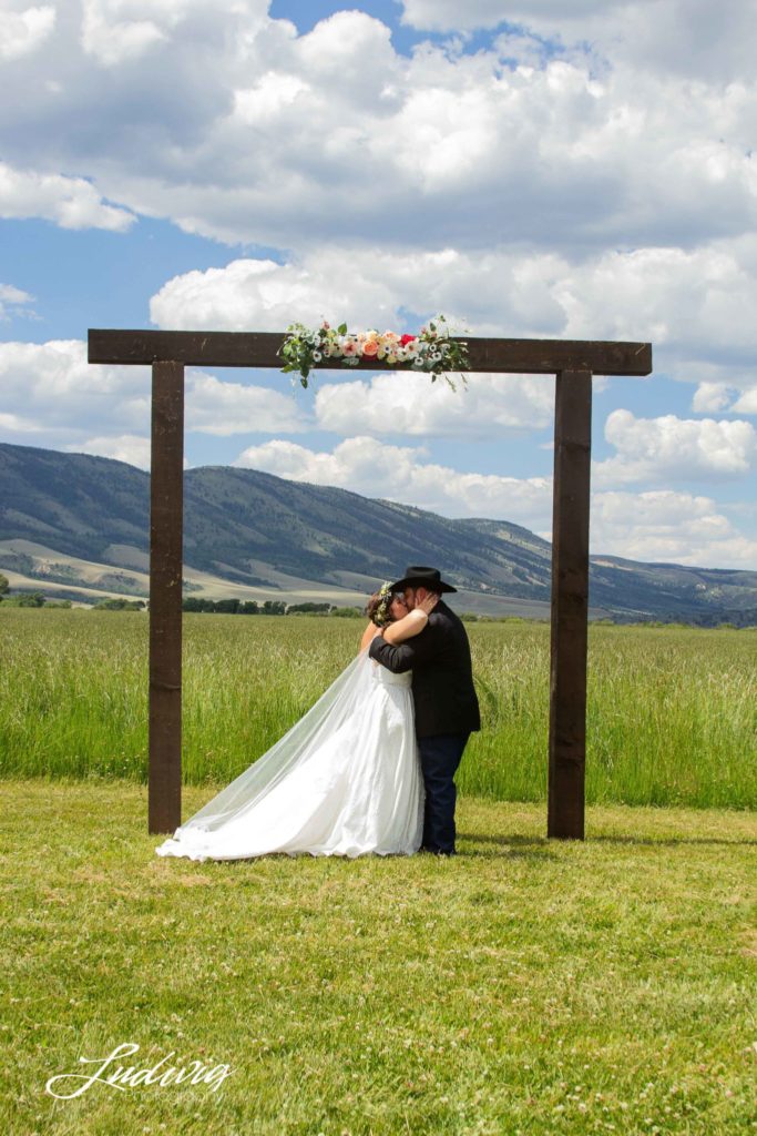 bride and groom kissing under wood arch with mountains in the background