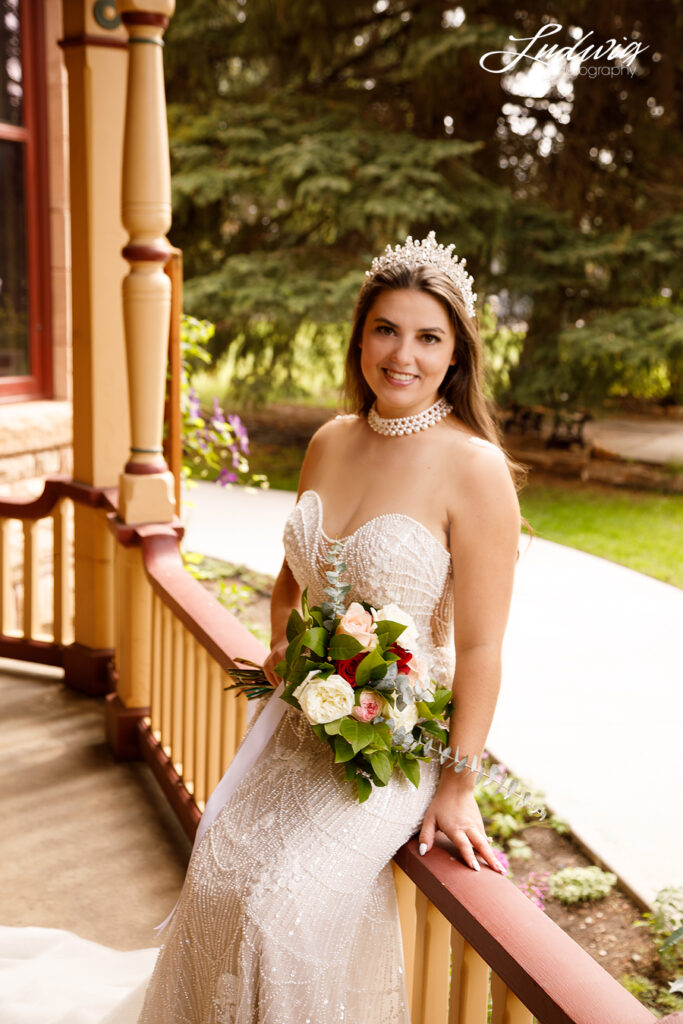 an outdoor portrait of a brunette bride in a wedding gown sitting on the railing of a porch with a garden in the background