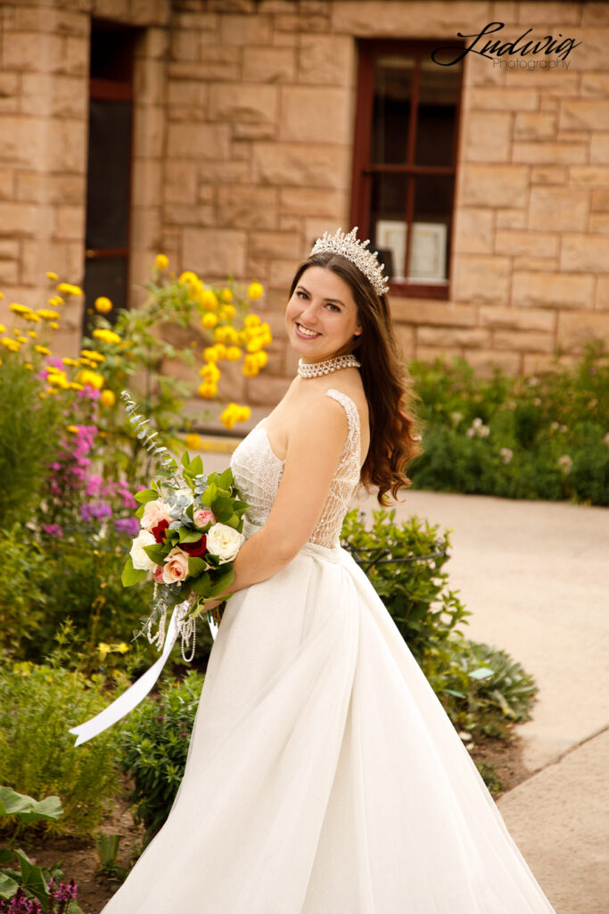 a beautiful brunette bride looks at the camera and smiles while clutching a rose bouquet in front of historic Ivinson Mansion