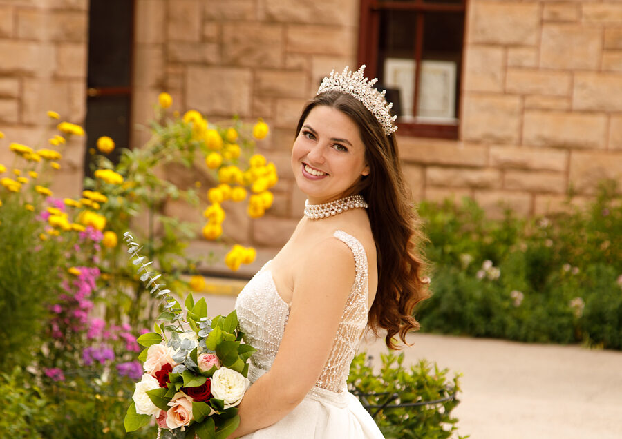 a beautiful brunette bride looks at the camera and smiles while clutching a rose bouquet in front of historic Ivinson Mansion