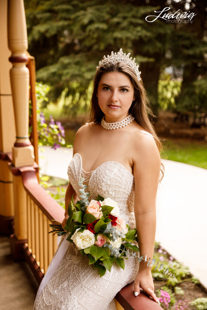 an outdoor portrait of a brunette bride in a wedding gown sitting on the railing of a porch with a garden in the background