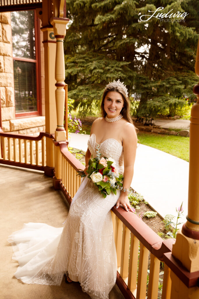 an outdoor portrait of a brunette bride in a wedding gown sitting on the railing of a porch with a garden in the background