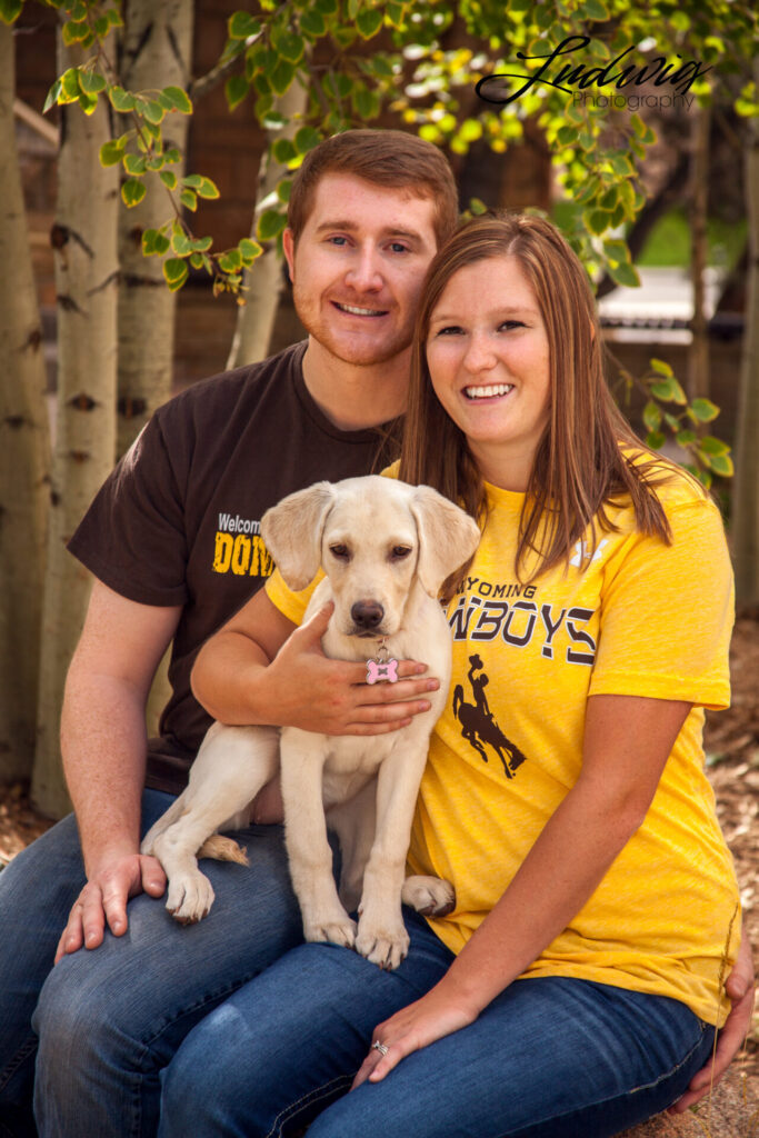 A couple sits together on the University of Wyoming campus smiling during a photoshoot with their puppy