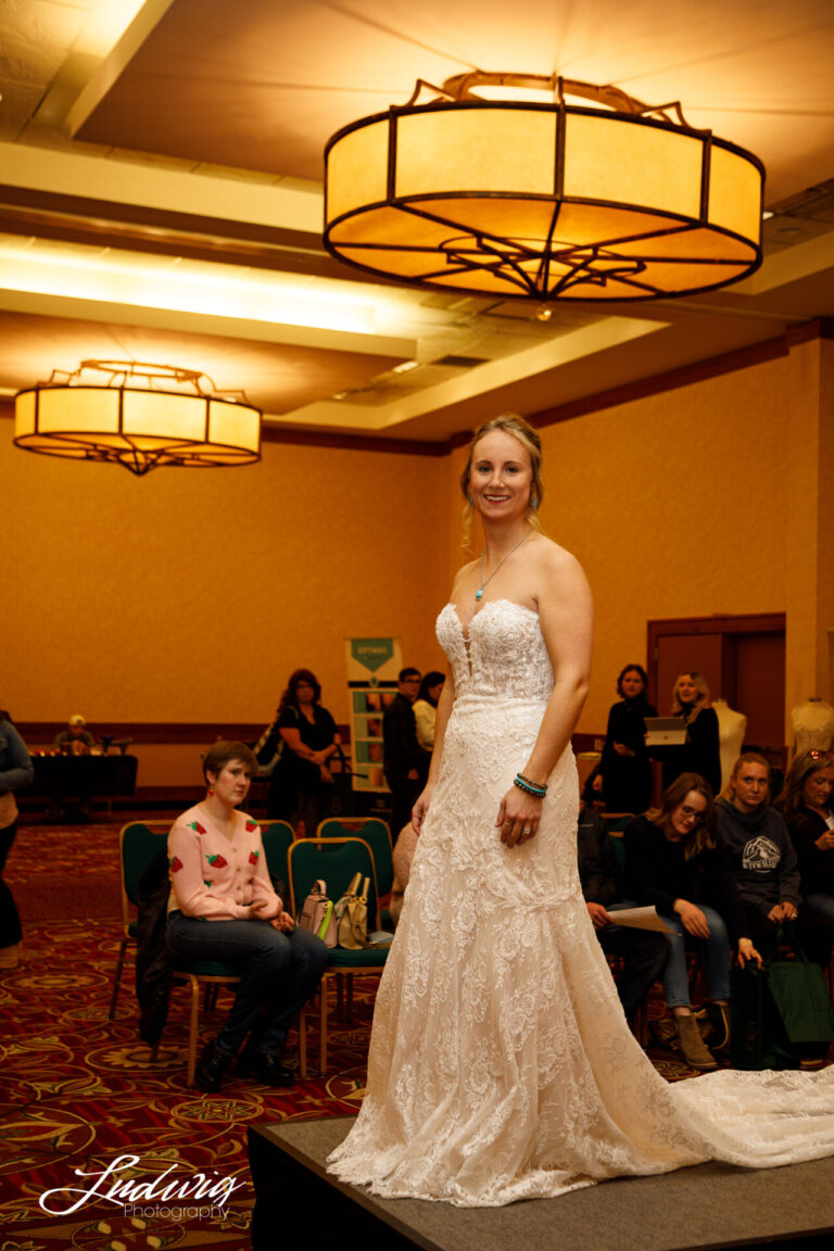 a young woman smiles at the camera on stage at fashion show wearing a lacy white wedding dress