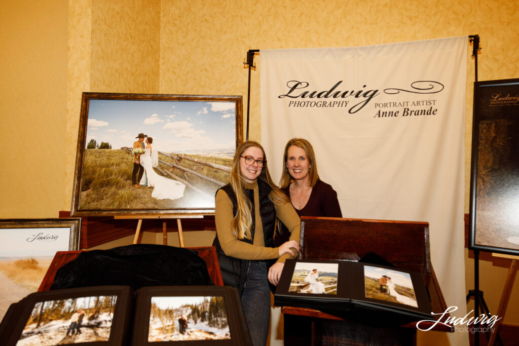 two women smile at the camera at a bridal expo with wall portraits of a bride and a cloth backdrop that says Ludwig Photography