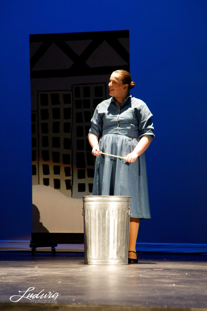 Girl standing in front of a trashcan with a wooden spoon, performing in Annie the musical.